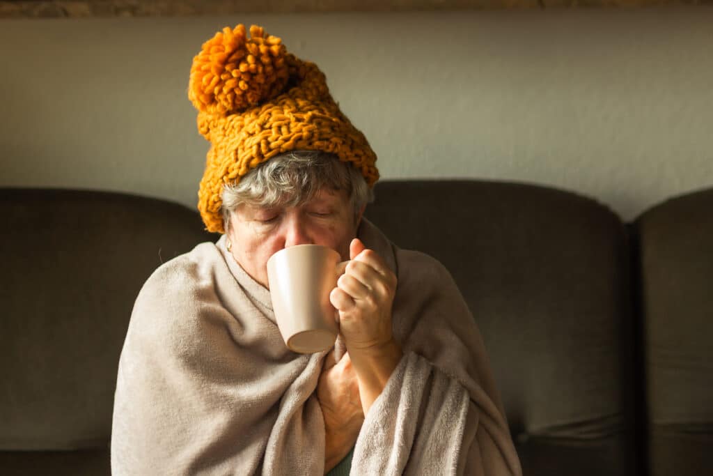 Elderly woman in knitted ginger cap wrapped in blanket and drinking hot cup of tea. Old grandma feeling cold at home with home heating trouble. Poverty and energy crisis in Europe. Cold symptoms.